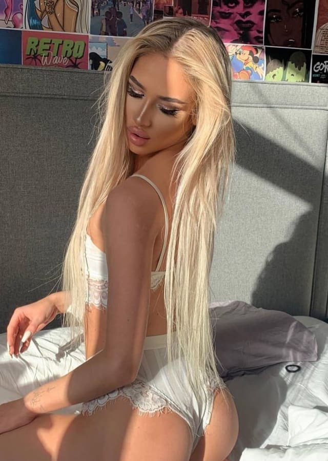 Long blonde hair, white lace lingerie, sitting on a bed with back turned, posters on the wall.