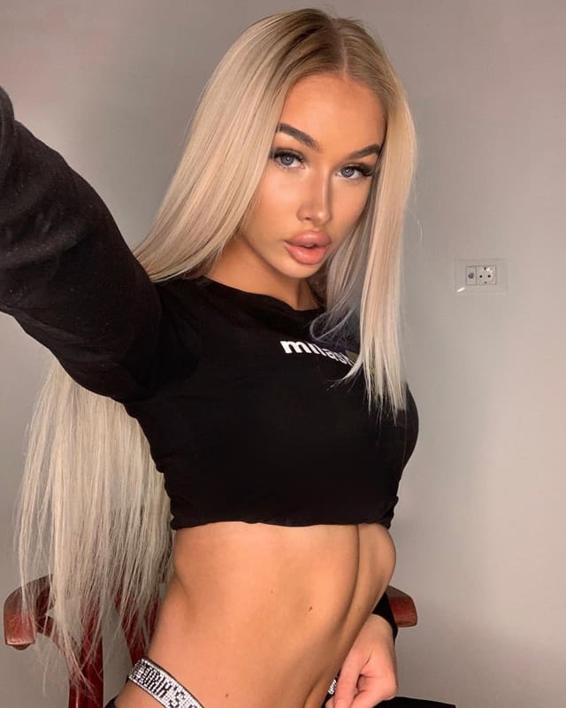 Polina with long blonde hair in a black crop top and sparkly belt, one arm raised against a plain wall.