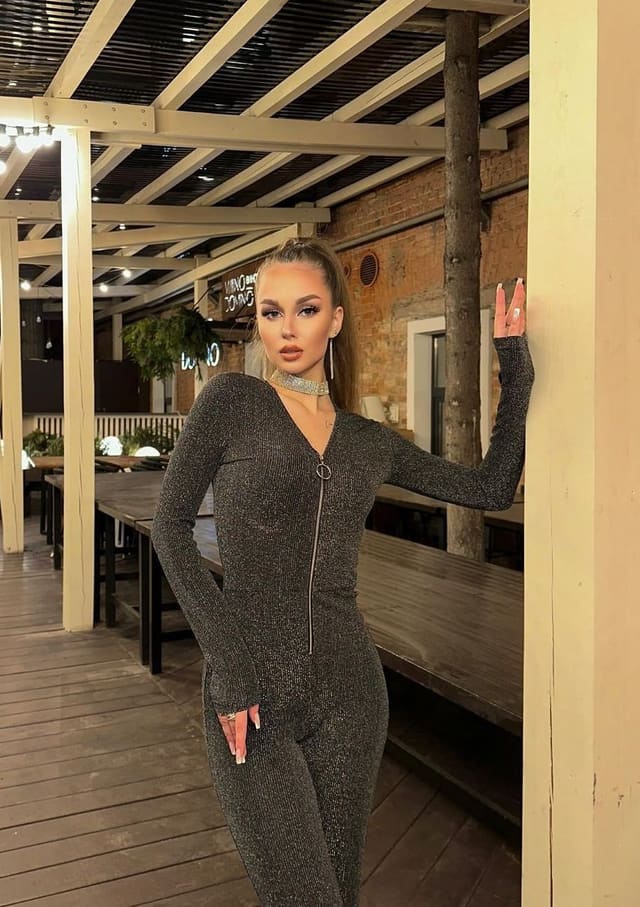 A woman in a sparkly gray jumpsuit stands on a wooden deck, hand on a post, hair tied back.