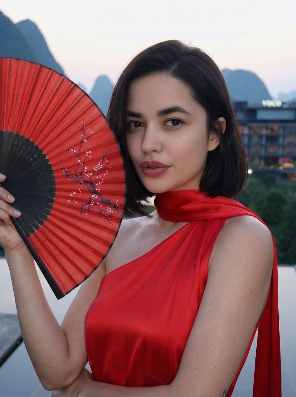 Lilian stands outdoors in a red one-shoulder dress holding a fan, with mountains and a building behind her.
