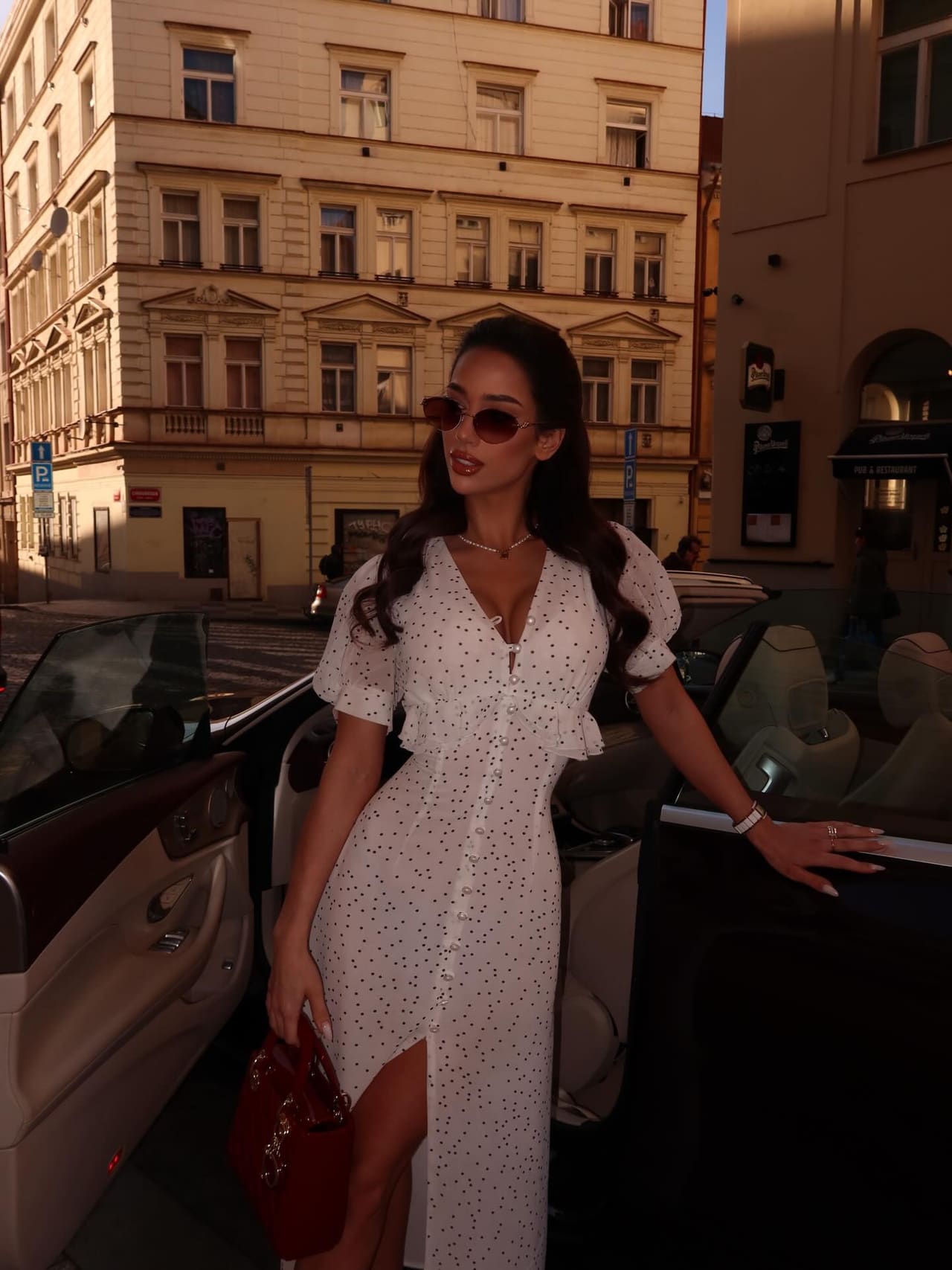 A woman with long dark wavy hair stands on a city street by an open car door in a white polka-dot midi dress with a thigh slit, holding a red handbag.