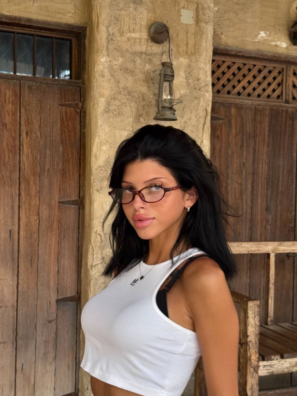 Lika in a white tank top stands before a rustic stone wall and wooden door.