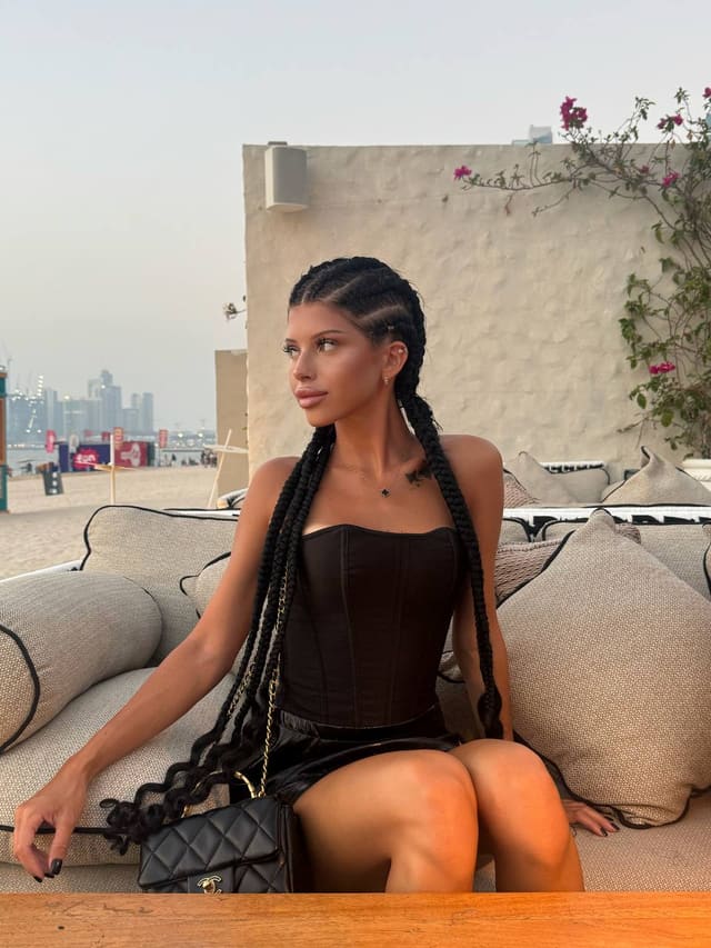 A woman with long black braided hair sits on an outdoor sofa, wearing a black strapless corset and skirt, with a bag on her lap near a beach.