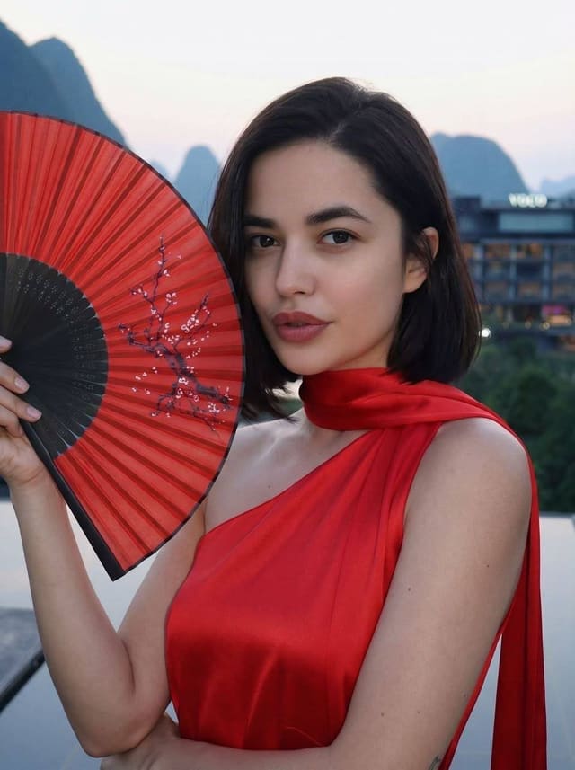 Lilian stands outdoors in a red one-shoulder dress, holding a red fan, with mountains and a building behind her.