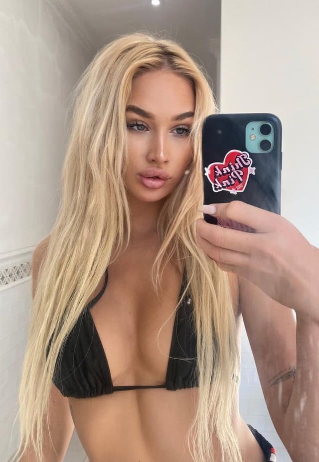 Long blonde hair, black bikini top, holding a phone with a heart sticker for a mirror selfie.