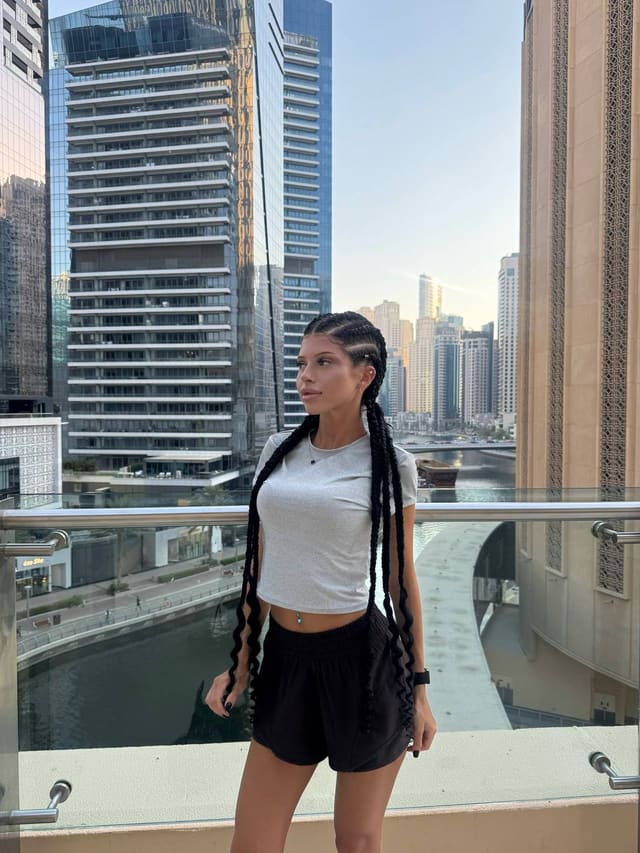 A woman with long braided hair stands on a balcony overlooking a city skyline, wearing a white short-sleeved crop top and black shorts.