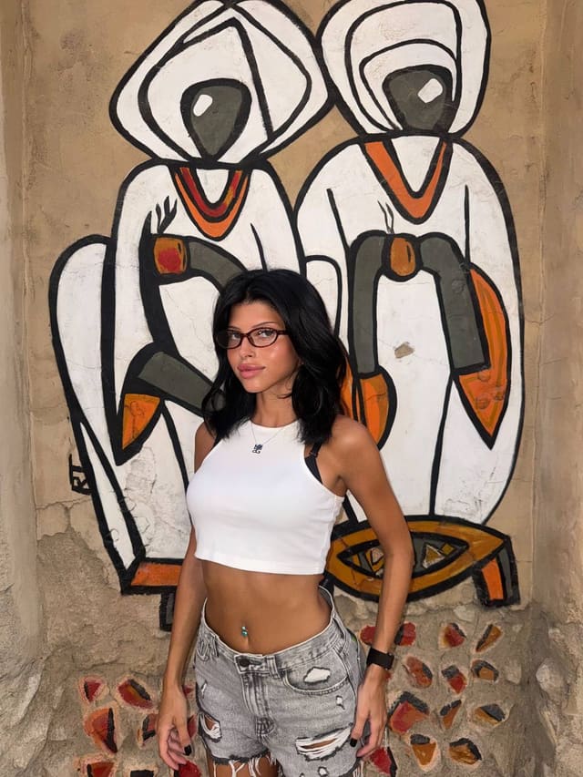 A woman with dark shoulder-length hair and glasses stands before a mural, wearing a white ribbed crop top and distressed denim shorts.