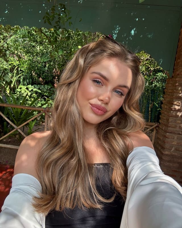 A woman with long wavy blonde hair and sunglasses on her head poses in a selfie, wearing a black top and white off-the-shoulder jacket against foliage and brick.