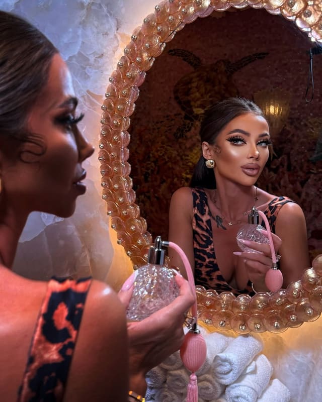 A woman with dark hair pulled back stands in profile holding a pink perfume bottle, her reflection in a decorative mirror showing her facing forward in a leopard-print top.