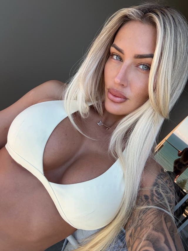 Anika with long blonde hair and a tattooed arm wears a white one-piece swimsuit with a deep neckline against a neutral background.