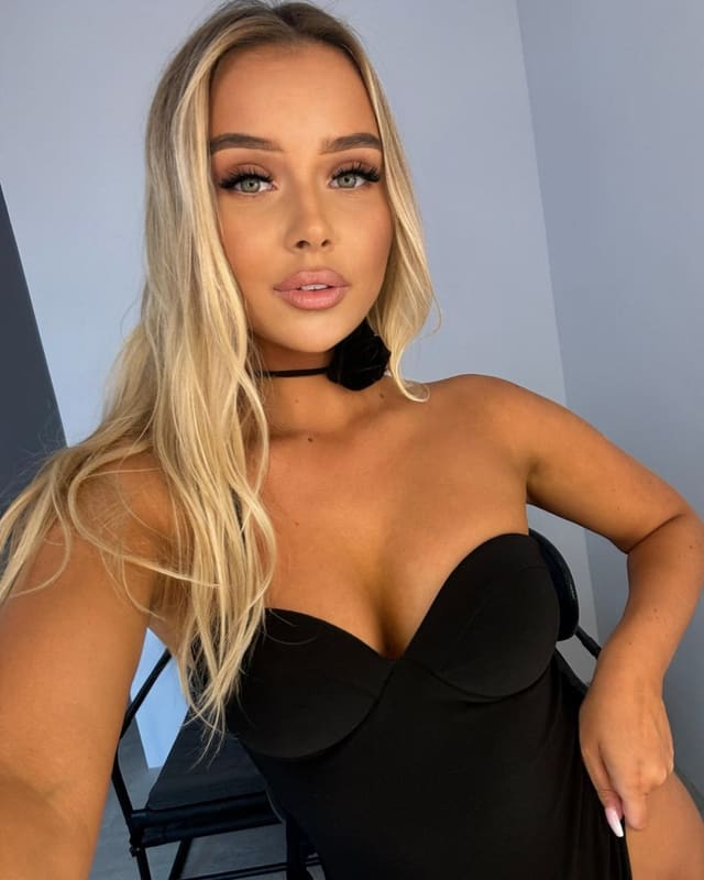 Daria with long blonde hair wears a black strapless dress and choker, posing with one hand on her hip against a light blue wall.