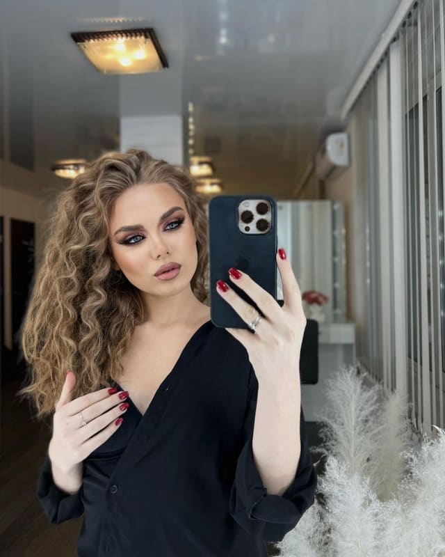 A woman with long curly blonde hair and red nails takes a mirror selfie in a black deep V-neck shirt, with vertical blinds and white plants in the background.