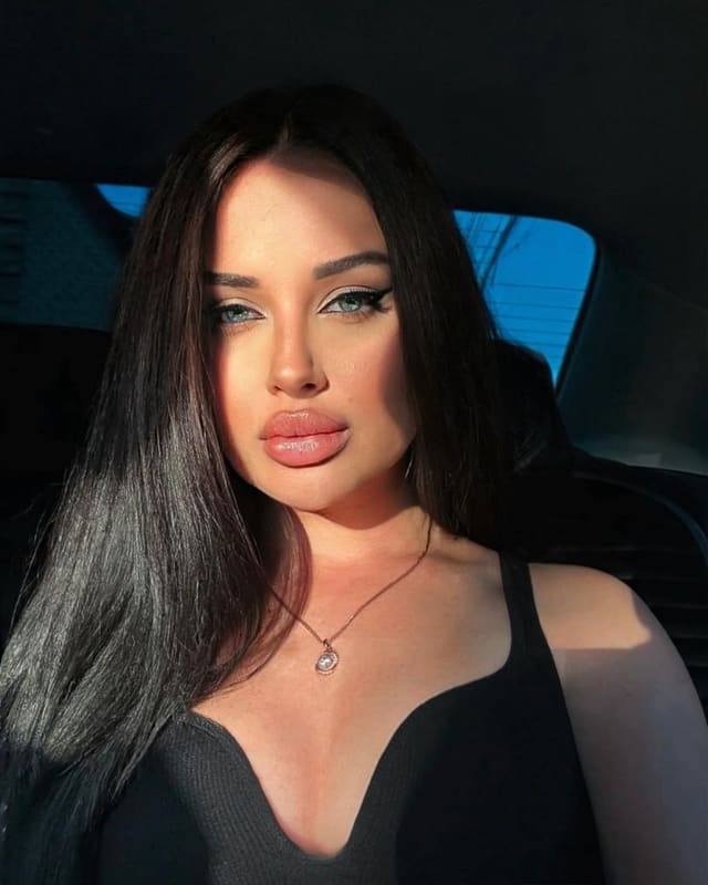 A person with long dark hair and winged eyeliner sits in a vehicle wearing a black tank top and necklace.