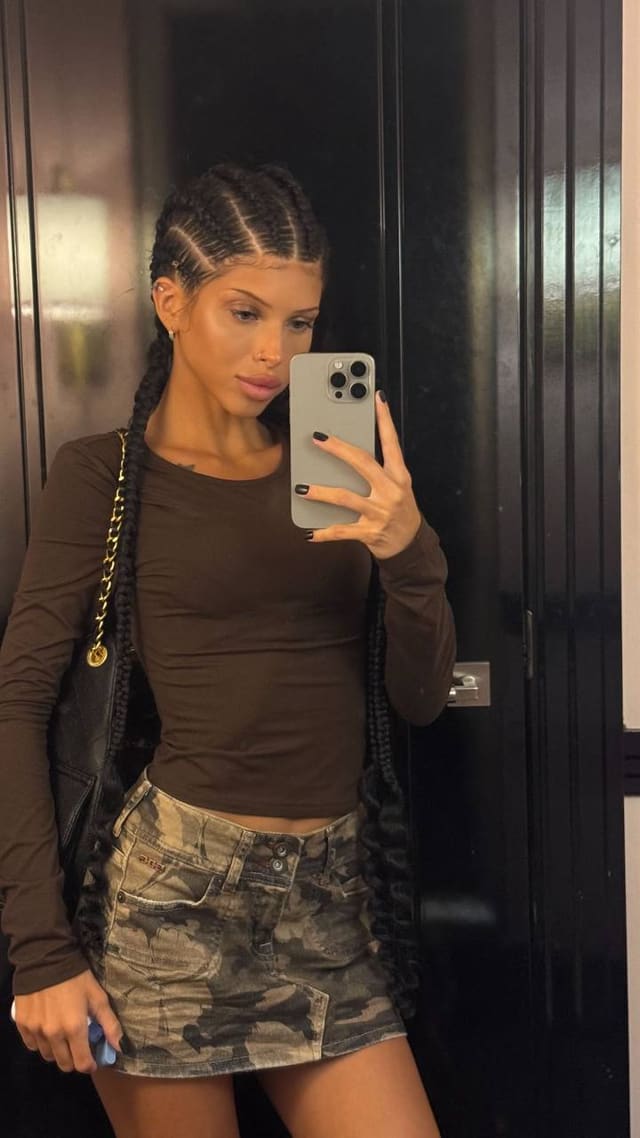 A person with long braided hair takes a mirror selfie, wearing a long-sleeved brown top and a camouflage mini skirt.