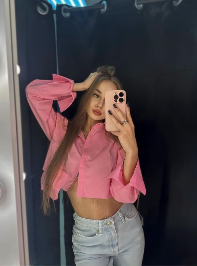 A person in a pink cropped shirt and jeans poses before a mirror, holding a phone.