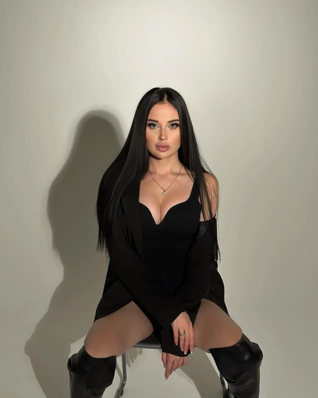 A person with long black hair sits on a chair against a plain background in a black dress and thigh-high boots.